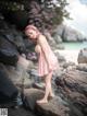 A woman in a pink dress standing on a rock by the water.